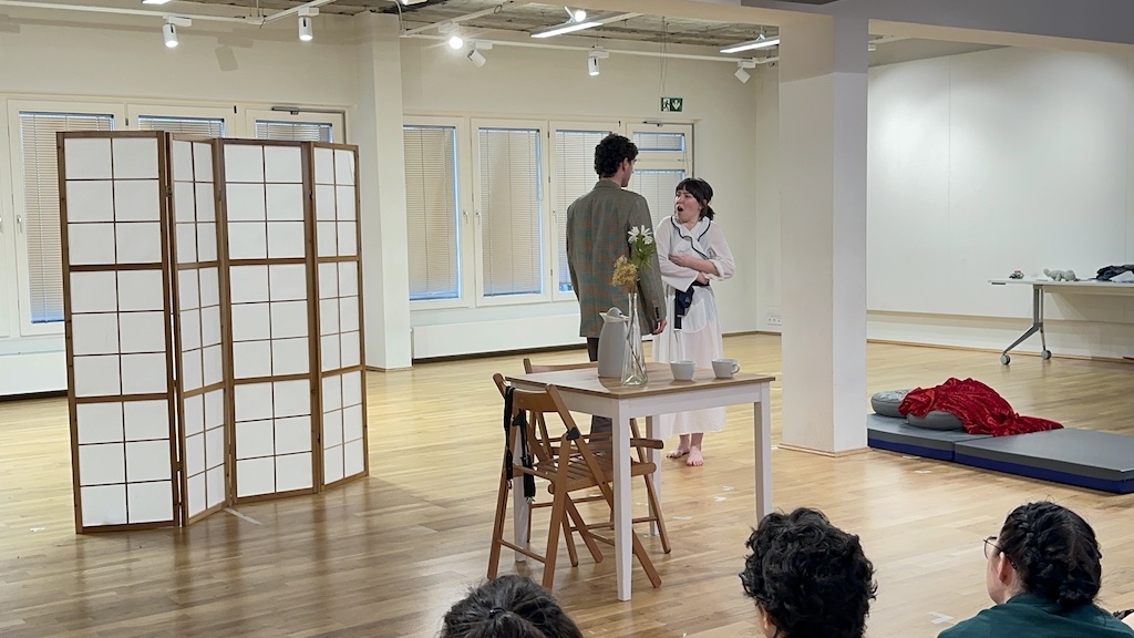 Two actors stand facing each other in mid-performance of 'Faith Hope Charity'. A table and chairs are set in front of the actors with a mattress and red blanket to their right.